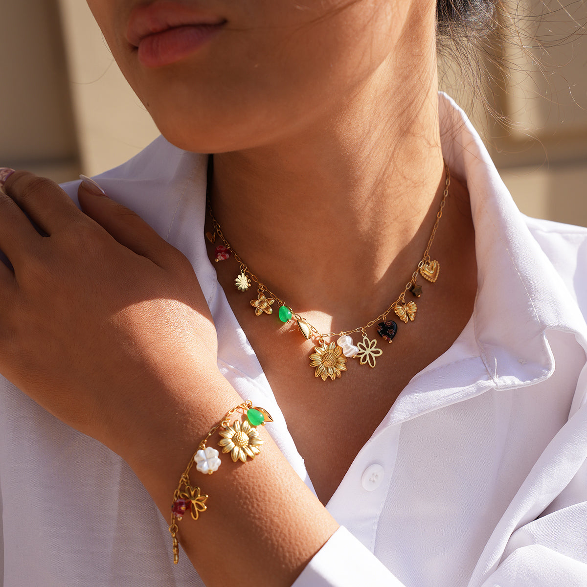 A close-up lifestyle shot of a model wearing a delicate gold necklace and matching bracelet. The jewelry features small gold charms, including flowers, hearts, and colorful gemstone accents in green, red, and white.